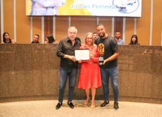 Cerimônia em plenário: uma mulher de vestido vermelho segura um certificado entre dois homens, um deles com um troféu nas mãos